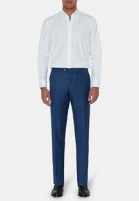 Man in een slim fit wit overhemd, blauwe pantalon en zwarte dresschoenen, staand tegen een effen witte achtergrond.