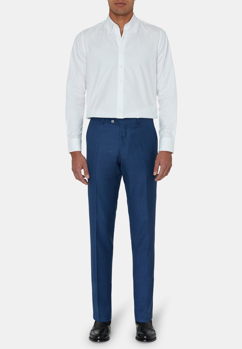 Man in een slim fit wit overhemd, blauwe pantalon en zwarte dresschoenen, staand tegen een effen witte achtergrond.