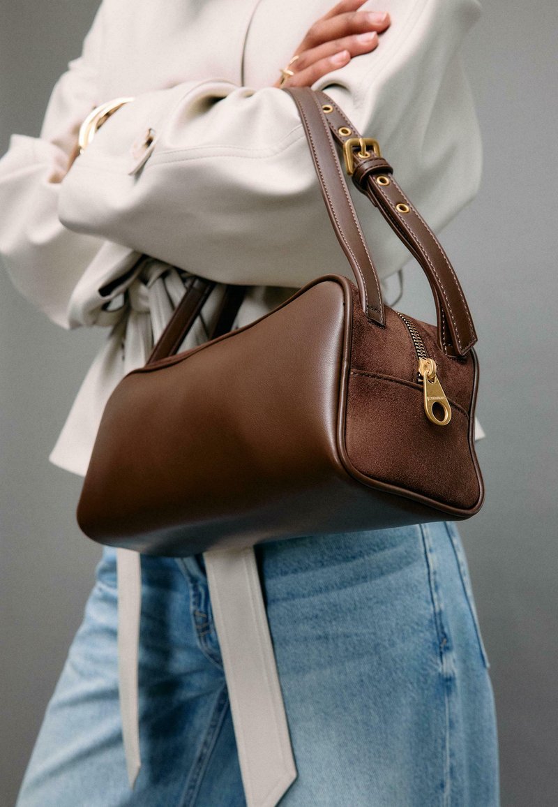 Personne portant une veste légère et un jean, tenant un sac à main en cuir marron avec une fermeture éclair dorée et une bandoulière ajustable, les bras croisés.