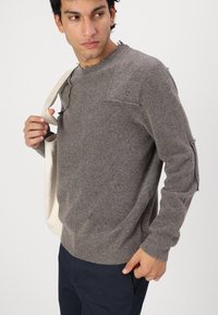 Grauer Strickpullover mit Rundhalsausschnitt, distressed Details und lockerer Passform. Kombiniert mit dunklen Hosen und einer cremefarbenen Tragetasche.