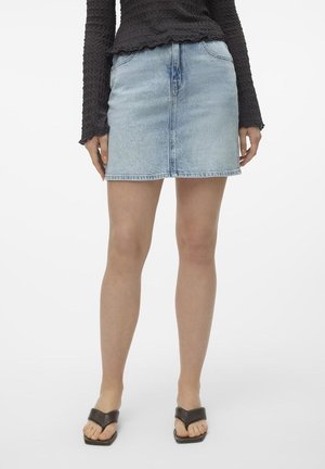 Personne portant une mini-jupe en denim bleu clair, un haut texturé à manches longues noir, et des sandales noires ouvertes à l'avant, debout sur fond blanc.