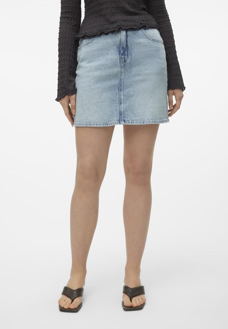Personne portant une mini-jupe en denim bleu clair, un haut texturé à manches longues noir, et des sandales noires ouvertes à l'avant, debout sur fond blanc.