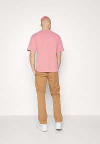 Man met kort roze haar, gekleed in een lichtroze t-shirt, beige broek en witte sneakers, staande tegenover een witte muur.