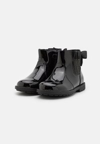 Bottines en cuir verni noir avec des panneaux élastiques sur les côtés, une semelle en caoutchouc plate et un accent de nœud décoratif à l'arrière.