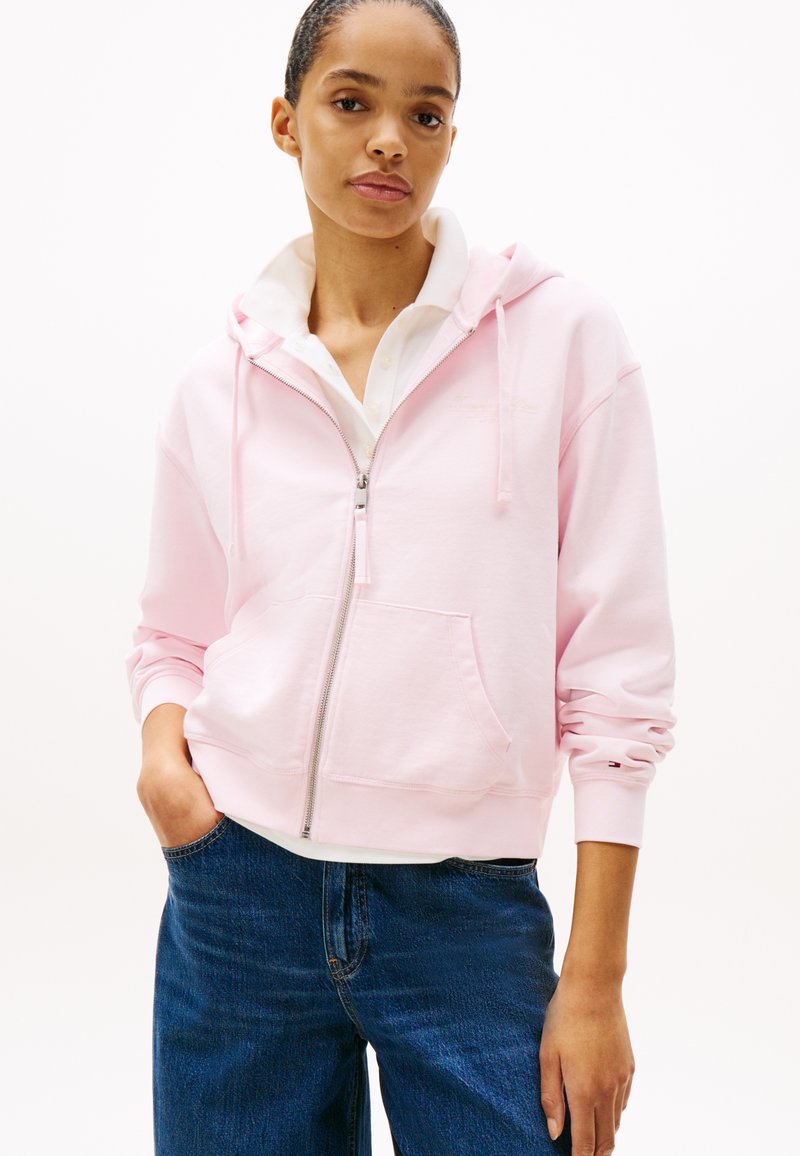 Felpa con zip rosa chiaro, dotata di tasca frontale, polsini a costine e una texture morbida, indossata sopra una camicia bianca con colletto e jeans blu.