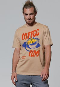 Beige katoenen T-shirt met een kleurrafische afbeelding van een croissant en een kop met de tekst "COFFEE CLUB" en "EVERYDAY ALL DAY" in oranje en blauw.