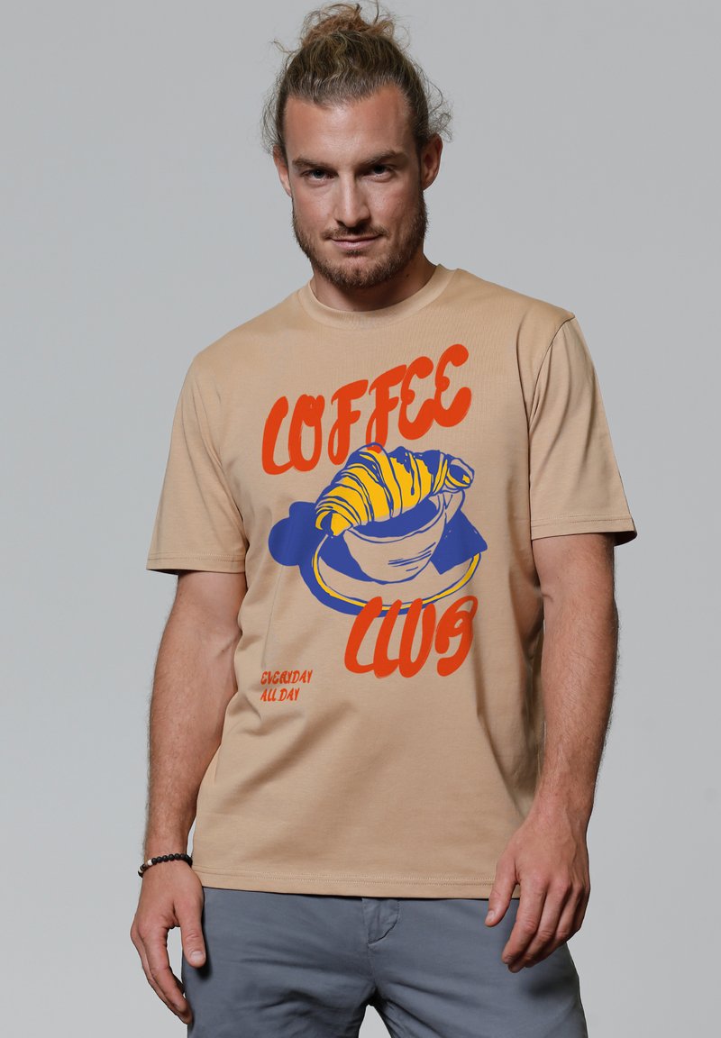 Beige katoenen T-shirt met een kleurrafische afbeelding van een croissant en een kop met de tekst "COFFEE CLUB" en "EVERYDAY ALL DAY" in oranje en blauw.