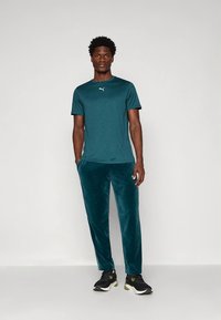 Kortermet sports skjorte i blågrønn med hvit logo, kombinert med blågrønne fløyelsjoggebukser og svarte treningssko. Enkel, sporty design.