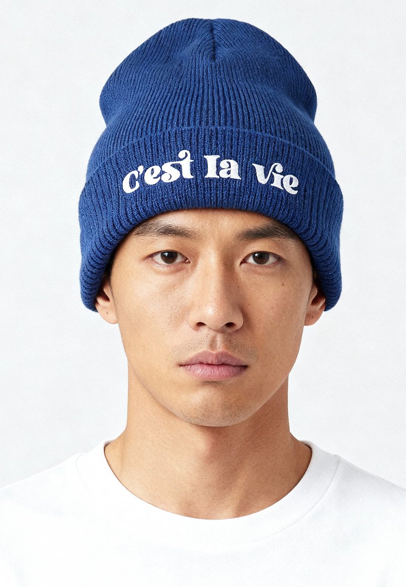 Bonnet côtelé bleu en matière tricotée, avec l'inscription « c'est la Vie » en texte cursif blanc sur le devant.