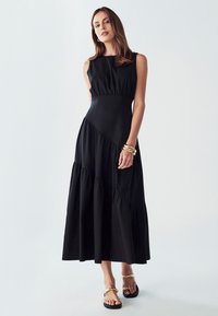 Črna brezrokavna obleka s prilagojenim telesom in slojevitim razširjenim robom. Narejena iz lahkega materiala z diskretnim sijajem in nabrano obdelavo.