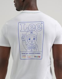 Un uomo indossa una camicia bianca con il logo blu "Lee" e un disegno colorato a numeri di "Buddy Lee" che mostra una figura che fa il segno di pace e una chiave di colori Crayola.