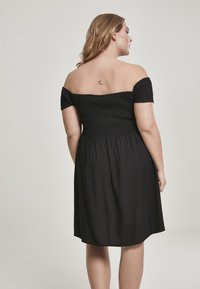 Svart off-shoulder-kjole med smocket liv og flytende skjørt. Glatt tekstur, knelengde, enkelt design og ingen synlige mønstre.