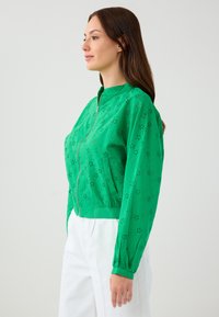 Groene ritsjack met bloempatroon van oogjes, lichtgewicht stof, cropped ontwerp en gathermanchetten. Draag over witte broeken.