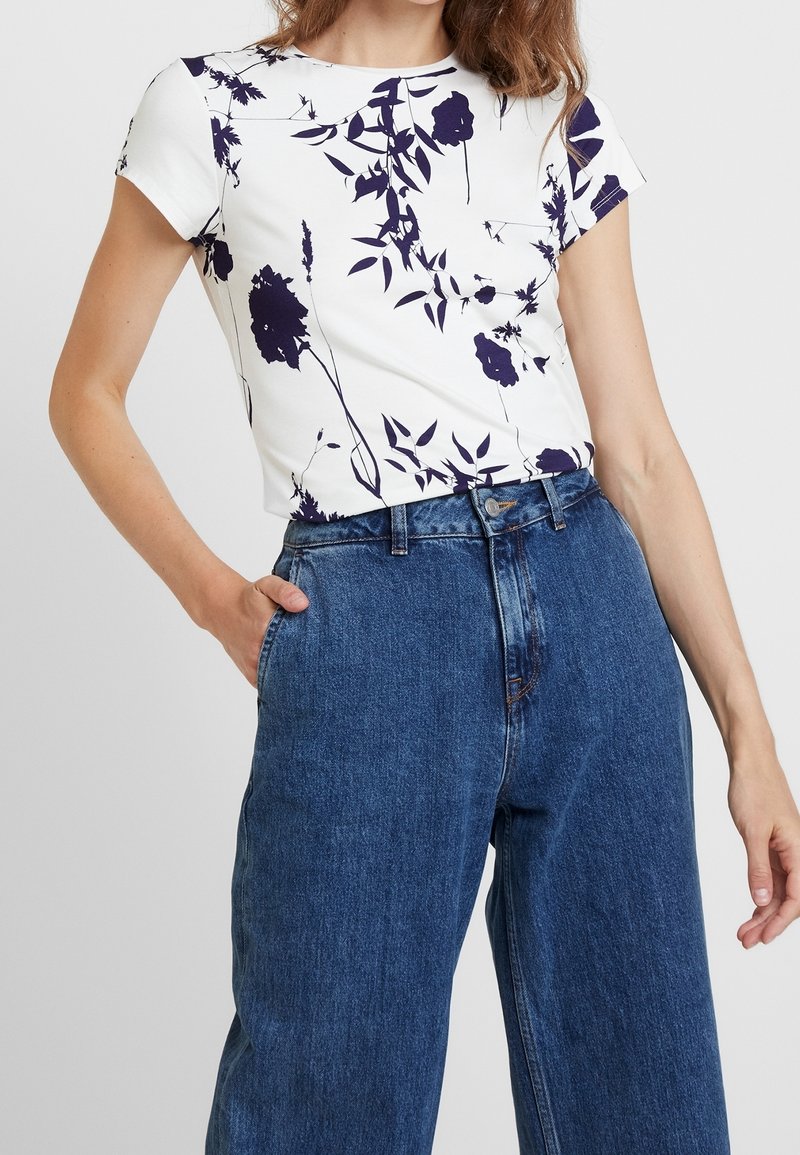Femme portant un haut blanc à manches courtes avec un imprimé floral foncé et un jean bleu ample taille haute, main dans la poche, sur un fond uni.