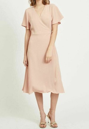Kobieta w jasnoróżowej sukience midi o kopertowym kroju z krótkimi, fruwającymi rękawami i złotych sandałkach na wysokim obcasie.