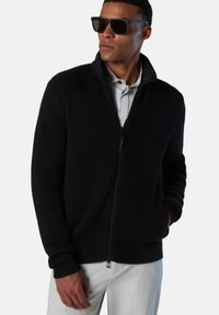 Schwarze, gerippte Zip-Jacke aus strukturiertem Stoff, mit hohem Kragen und taillierten Bündchen, kombiniert mit einem hellfarbenen Poloshirt darunter.