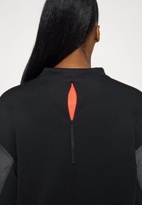 Top atletico nero con una ventilazione sul retro e un accento rosso, chiusura con zip e maniche grigie texturizzate. Adatto per l'abbigliamento sportivo.