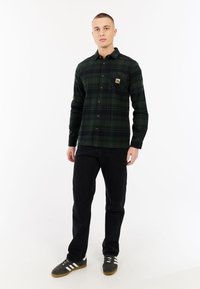 Man in een donker groen en zwart geruit overhemd met knopen, zwarte broek en grijze sneakers met witte strepen, staande tegen een effen witte achtergrond.