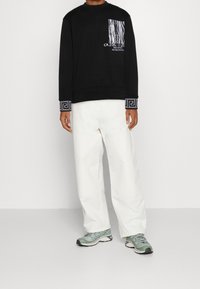 Felpa nera con stampa grafica e tasca testurizzata, dotata di polsini a motivi. Abbinata a pantaloni bianchi larghi e sneakers verdi.