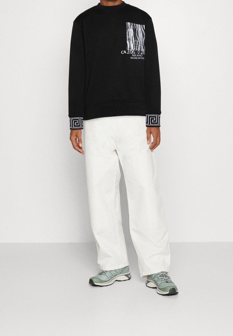 Felpa nera con stampa grafica e tasca testurizzata, dotata di polsini a motivi. Abbinata a pantaloni bianchi larghi e sneakers verdi.