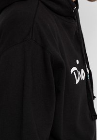 Detailní záběr na osobu, která má na sobě černou mikinu s kapucí, na hrudi s částečným bílým textem a černými šňůrkami na kapuci.