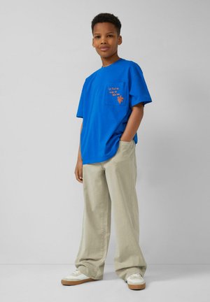 Chłopiec stojący z jedną ręką w kieszeni, ubrany w jasnoniebieską koszulkę z pomarańczowym napisem, beżowe spodnie z szerokimi nogawkami i białe trampki.
