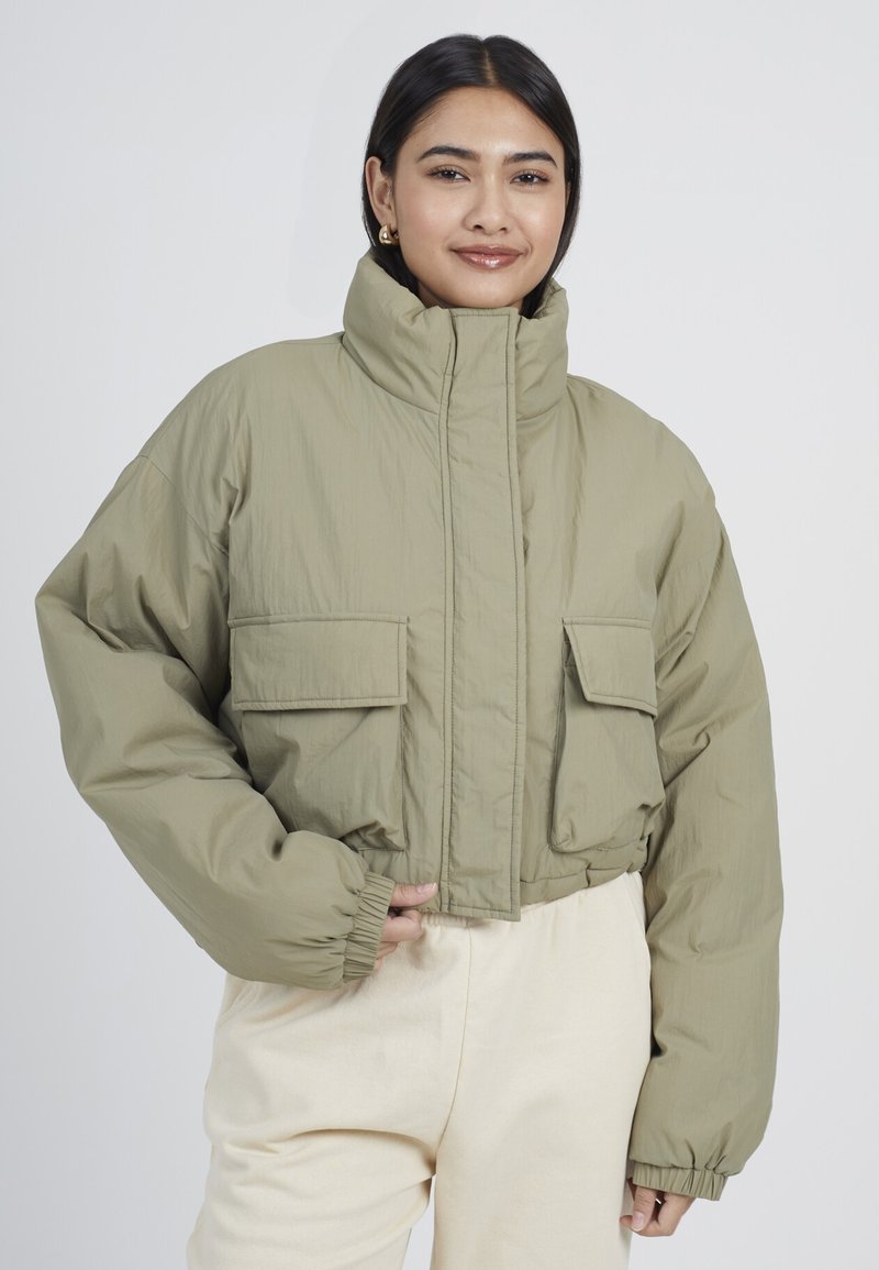 Chaqueta acolchada de color verde oliva recortada, con cuello alto, dos bolsillos frontales, puños elásticos y textura de tela suave y ligera.