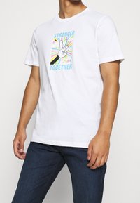 Bílý bavlněný tričko s barevným grafickým motivem znaku míru, textem „SILNĚJŠÍ SPOLEČNĚ“ a „PUMA“, umístěným na pozadí slunečních paprsků.