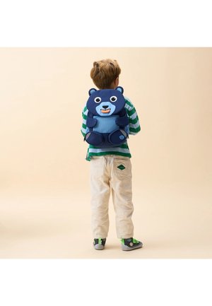 Child wearing green and white striped shirt, beige pants, blue bear-shaped backpack, and grey shoes with green laces standing against beige background.