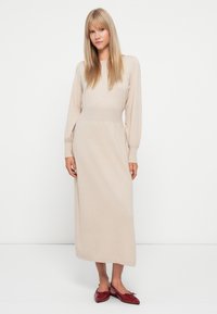 Abito lungo a maniche lunghe beige in maglia con corpino aderente e gonna morbida, abbinato a scarpe basse rosse. Tessuto liscio, design minimalista.