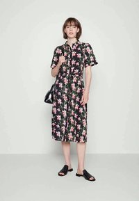 Jovem mulher em pé, vestindo um vestido midi preto com flores rosas, algumas sandálias pretas abertas nas costas e segurando uma mala preta no ombro.