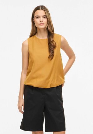 Kvinde med langt brunt hår iført en sennepsfarvet ærmeløs top og sorte knælange shorts, stående med en hånd bag ryggen.