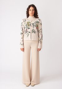 Gebreide cardigan in crème met bloemendessin; hoge kraag, sluiting aan de voorkant; gecombineerd met beige broek met wijde pijpen en beige schoenen.
