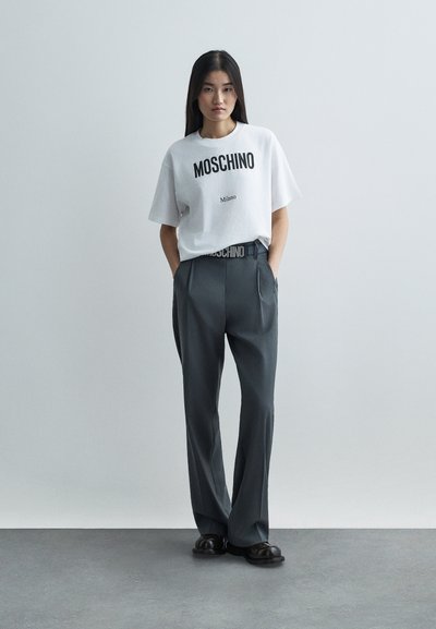 Donna con lunghi capelli scuri che indossa una maglietta bianca Moschino Milano infilata in pantaloni grigi a pieghe e scarpe nere, in piedi con le mani in tasca.