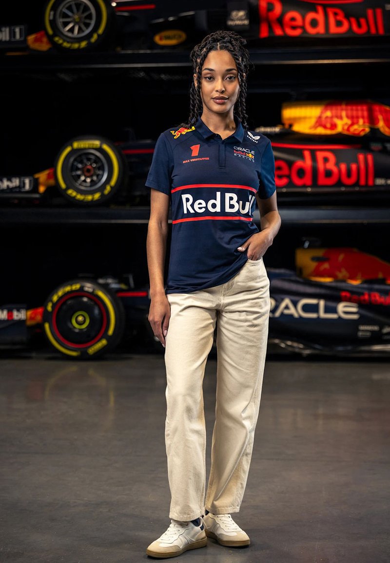 Granatowa koszulka polo z czerwonymi i białymi akcentami, z logo i numerem. W zestawieniu z kremowymi spodniami i białymi trampkami, na tle wyścigowych samochodów.