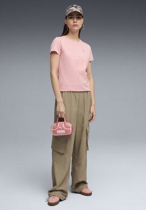 Femme portant un t-shirt rose, un pantalon cargo beige, une casquette camouflage, des chaussures roses et tenant un petit sac à main rose Puma sur un fond gris uni.