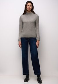 Frau steht und trägt einen grauen Rollkragenpullover, dunkelblaue Jeans mit weitem Bein und schwarze Stiefeletten vor einem schlichten, hellen Hintergrund.