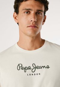 Jeune homme aux cheveux courts et bouclés portant un T-shirt blanc Pepe Jeans London sur un fond uni.