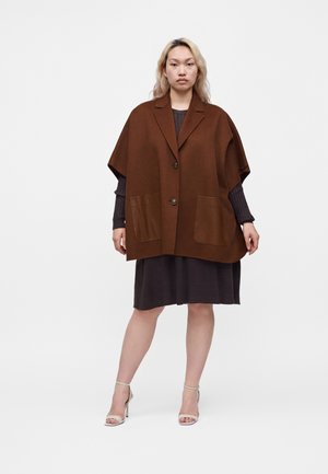 Femme debout face à l'objectif, portant une veste cape marron oversize avec de grandes poches frontales, une robe noire et des sandales beiges à talons hauts.
