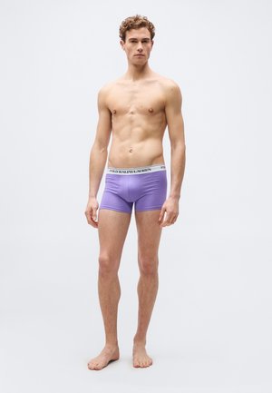 Jeune homme debout pieds nus portant un boxer violet avec une ceinture blanche à logo, sur un fond clair uni.