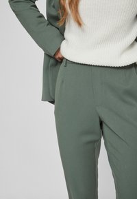 Roheline kohandatud pükskostüüm ja valge kudum; püksid on valmistatud siledast kangast, sirge lõikega ja neil on peened esipleatsid.