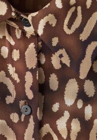 Camisa de tela marrón con manchas irregulares en óvalo y circular beige, con botones negros y cuello doblado.