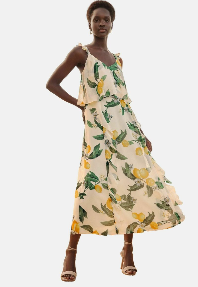 Maxime dress in cream with a lemon and green leaf floral print. Features thin shoulder straps, ruffled layers, and a flowing silhouette.