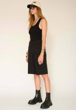 Débardeur noir, short noir ajusté, bottes noires à semelle épaisse et casquette beige avec lettrage noir. Les accessoires incluent des bracelets et des bagues.