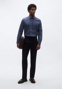 Mėlyna marškiniai mygtukais ir tamsios kelnės, derinamos su ruda odine laiškininko krepše; modelis stovi ant paprasto fono.