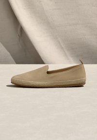 Μπεζ casual παπούτσι slip-on με λείο πάνω μέρος, πλεκτή σόλα από σχοινί και μικρό ταμπ, τοποθετημένο σε ουδέτερο ύφασμα και φόντο επιφάνειας.