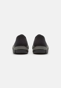 Black slip-on shoes with a textured upper, featuring a rubber sole and grey accents at the heel and midsole. Minimalist design.