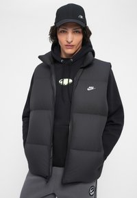 Jovem vestido com um boné preto da Nike, um colete acolchoado preto, um moletom preto e calças de moletom cinzentas da Nike, de pé com as mãos nos bolsos contra um fundo branco.