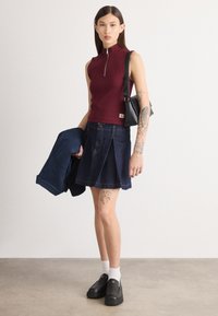 Haut sans manches bordeaux avec col zippé, associé à une jupe plissée en denim foncé. Chaussures noires à enfiler et petit sac à main noir.