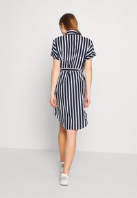 Abito camicia a righe blu navy e bianche con maniche corte, vita con cintura e taglio dritto. Abbinato a sneaker bianche. Tessuto dalla struttura liscia.